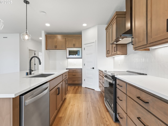 kitchen featuring a sink, wall chimney range hood, appliances with stainless steel finishes, light wood-style floors, and brown cabinets