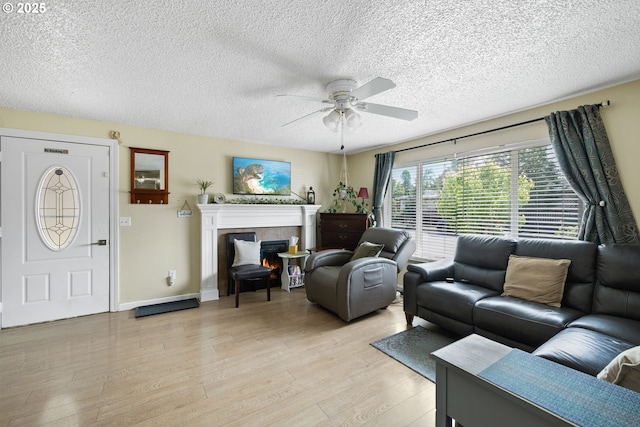 living room with ceiling fan, light wood-style flooring, a textured ceiling, a tiled fireplace, and baseboards