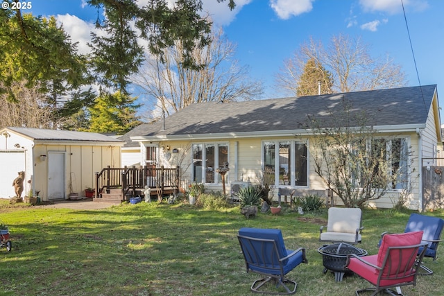 rear view of property with an outbuilding, a shingled roof, a lawn, an outdoor fire pit, and a wooden deck