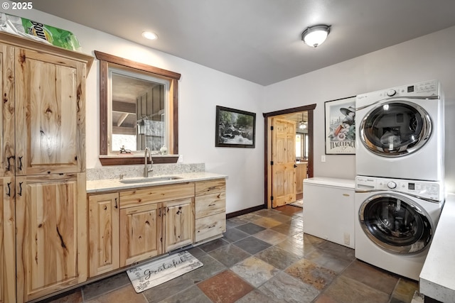 clothes washing area featuring stacked washer and dryer, stone tile flooring, cabinet space, a sink, and baseboards