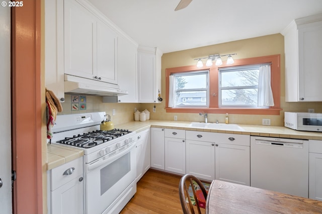 kitchen featuring tile counters, under cabinet range hood, white cabinets, white appliances, and a sink