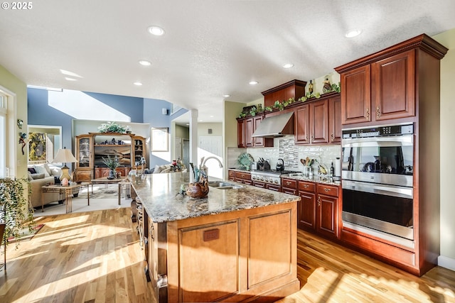 kitchen with stone countertops, sink, stainless steel double oven, a center island with sink, and wall chimney exhaust hood