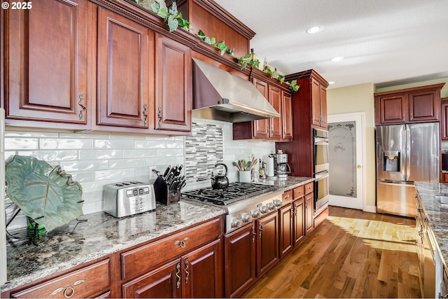 kitchen with stone counters, stainless steel appliances, hardwood / wood-style floors, and wall chimney range hood