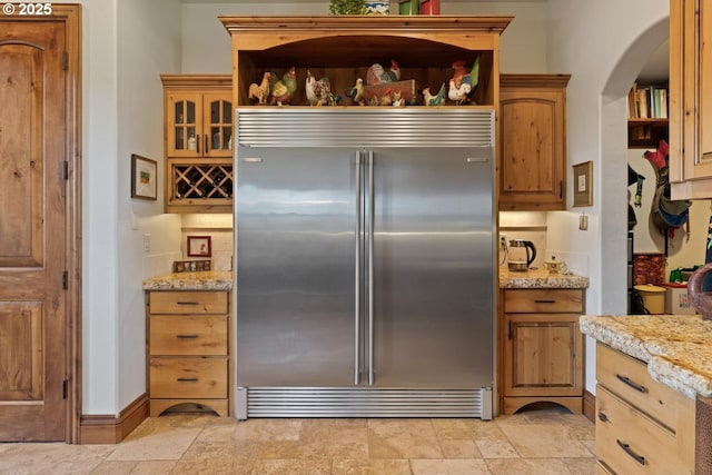 kitchen featuring open shelves, stone finish floor, stainless steel built in fridge, light stone counters, and arched walkways