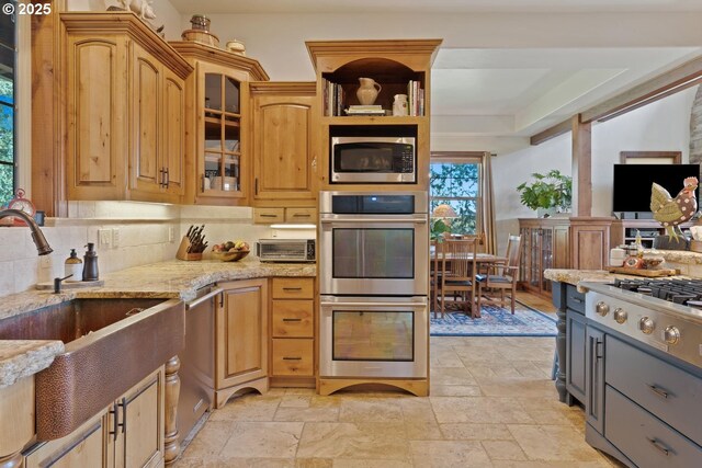 kitchen with stone tile flooring, light stone countertops, glass insert cabinets, a sink, and stainless steel appliances