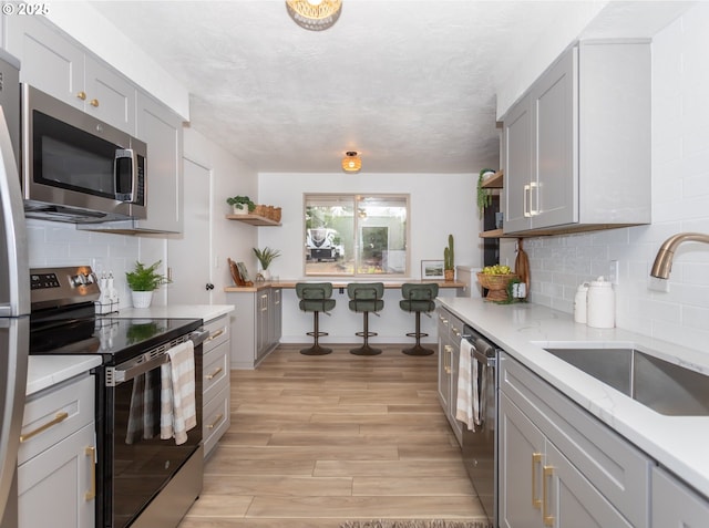 kitchen featuring open shelves, gray cabinets, a sink, stainless steel appliances, and light wood-type flooring