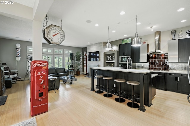 10155 SW Morrison St, Portland, OR 97225 - kitchen with wall chimney exhaust hood, dark cabinets, light wood-style floors, decorative backsplash, and a kitchen bar