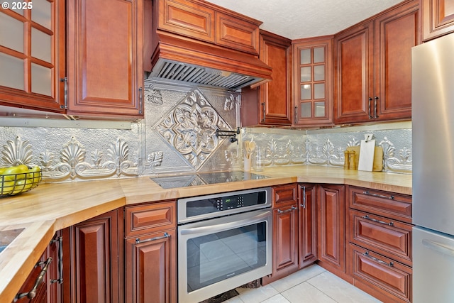 kitchen featuring appliances with stainless steel finishes, butcher block countertops, custom range hood, light tile patterned floors, and glass insert cabinets