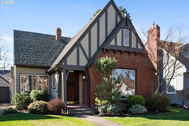 tudor house with brick siding, roof with shingles, a chimney, stucco siding, and a front lawn