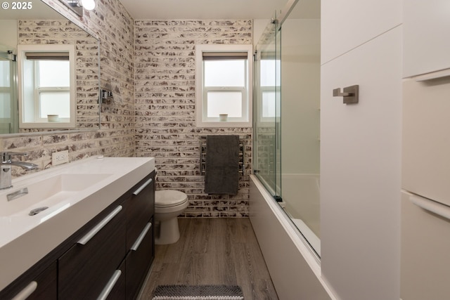 full bathroom with brick wall, vanity, bath / shower combo with glass door, toilet, and hardwood / wood-style flooring