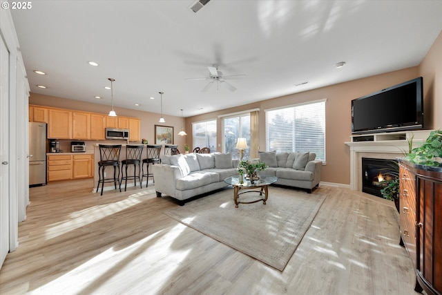 living room featuring visible vents, light wood-style flooring, a ceiling fan, and a glass covered fireplace