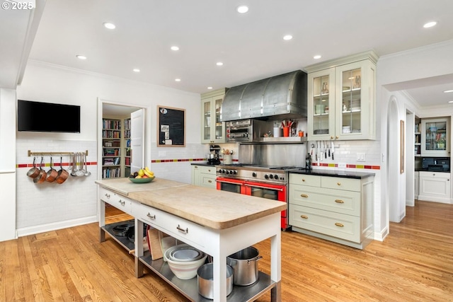 kitchen with wall chimney range hood, double oven range, open shelves, light wood-type flooring, and ornamental molding