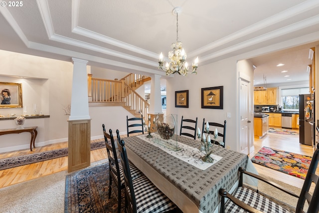 dining space featuring a raised ceiling, ornate columns, ornamental molding, light wood-type flooring, and a notable chandelier