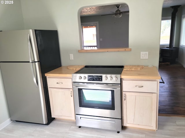 kitchen with a ceiling fan, light brown cabinetry, appliances with stainless steel finishes, a wood stove, and light wood-style flooring