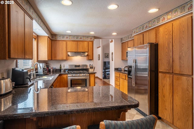 kitchen featuring a sink, under cabinet range hood, stainless steel appliances, a peninsula, and brown cabinetry