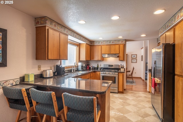 kitchen featuring a peninsula, a sink, stainless steel appliances, under cabinet range hood, and dark countertops