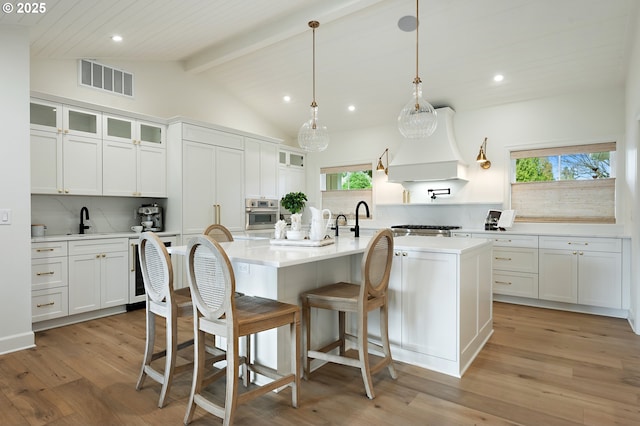 kitchen featuring custom range hood, a kitchen island with sink, backsplash, light countertops, and white cabinets
