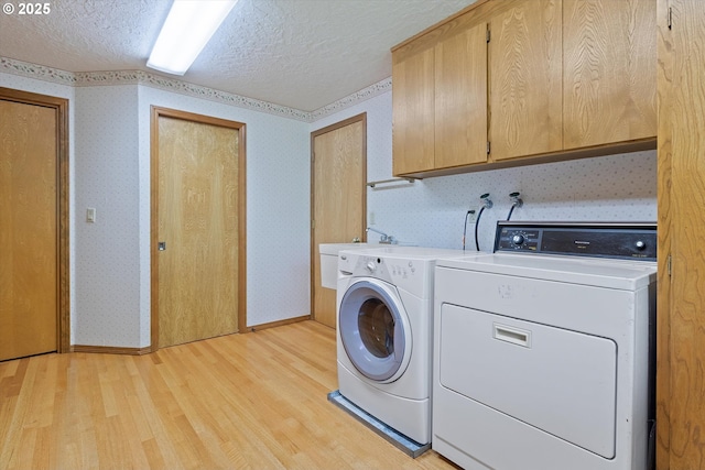 clothes washing area featuring a textured ceiling, light wood-style flooring, separate washer and dryer, cabinet space, and wallpapered walls