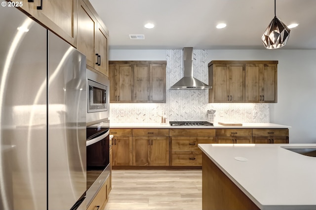 kitchen with wall chimney range hood, stainless steel appliances, tasteful backsplash, light hardwood / wood-style floors, and decorative light fixtures