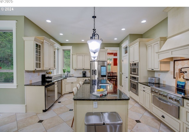 kitchen with cream cabinetry, a warming drawer, a center island with sink, stainless steel appliances, and glass insert cabinets