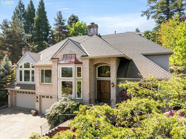 view of front facade with roof with shingles, a chimney, brick siding, an attached garage, and driveway