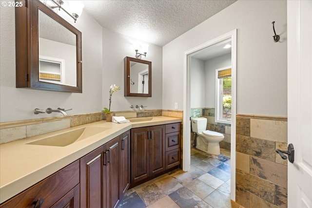 bathroom featuring double vanity, a textured ceiling, a sink, toilet, and stone tile flooring