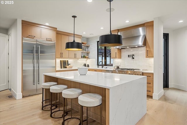 kitchen with appliances with stainless steel finishes, a kitchen island, decorative light fixtures, wall chimney range hood, and light hardwood / wood-style floors