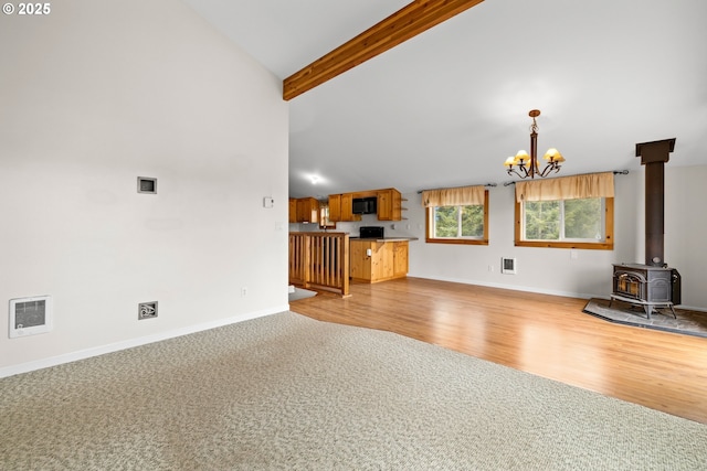 unfurnished living room featuring a wood stove, a chandelier, light colored carpet, beam ceiling, and baseboards