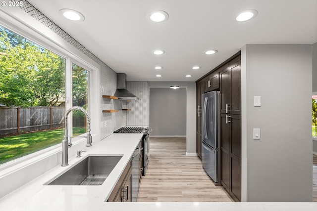 kitchen with appliances with stainless steel finishes, a sink, light wood finished floors, recessed lighting, and wall chimney exhaust hood