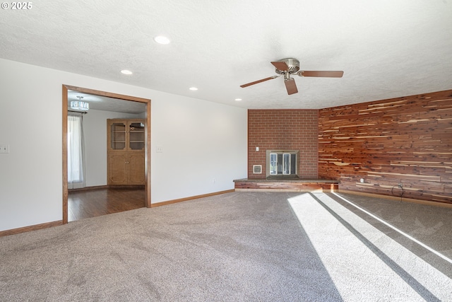 unfurnished living room with wood walls, ceiling fan, dark carpet, a brick fireplace, and a textured ceiling