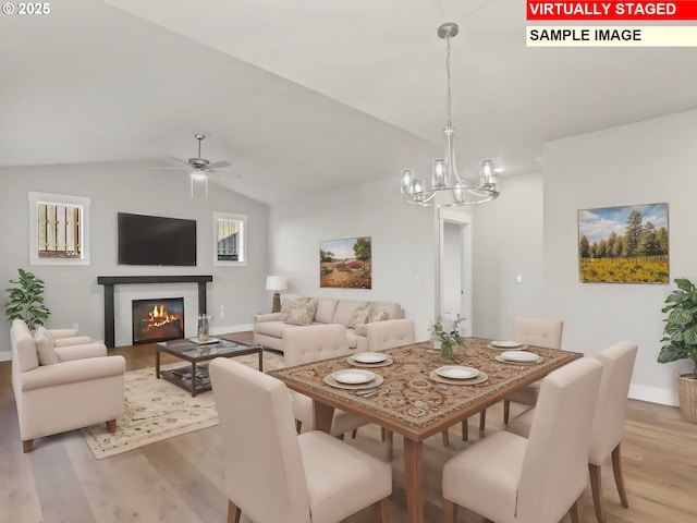 dining space featuring a glass covered fireplace, light wood-type flooring, baseboards, lofted ceiling, and ceiling fan with notable chandelier