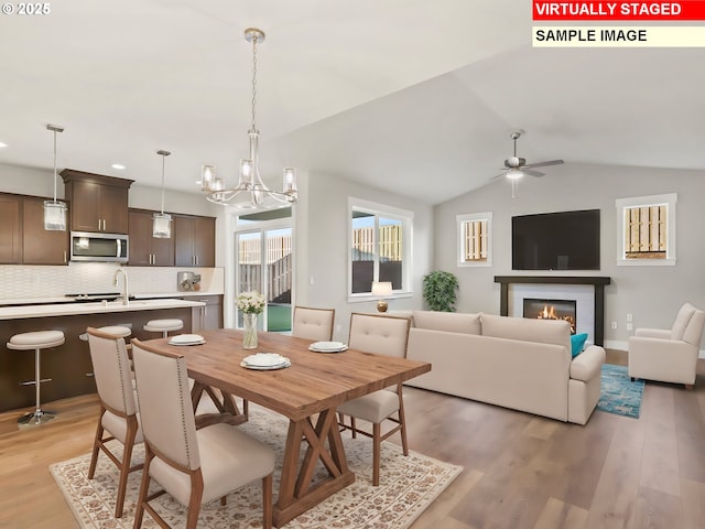 dining space featuring light wood-type flooring, a glass covered fireplace, vaulted ceiling, and ceiling fan with notable chandelier