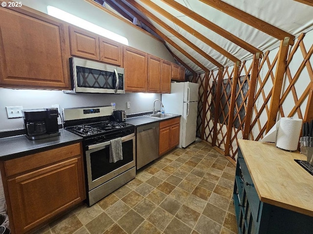 kitchen with stainless steel appliances, stone finish flooring, a sink, vaulted ceiling, and brown cabinetry