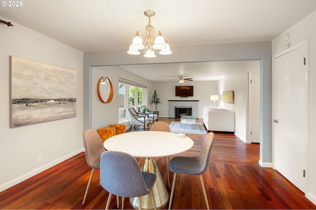 dining space featuring a chandelier, dark wood-style flooring, a fireplace, baseboards, and a ceiling fan