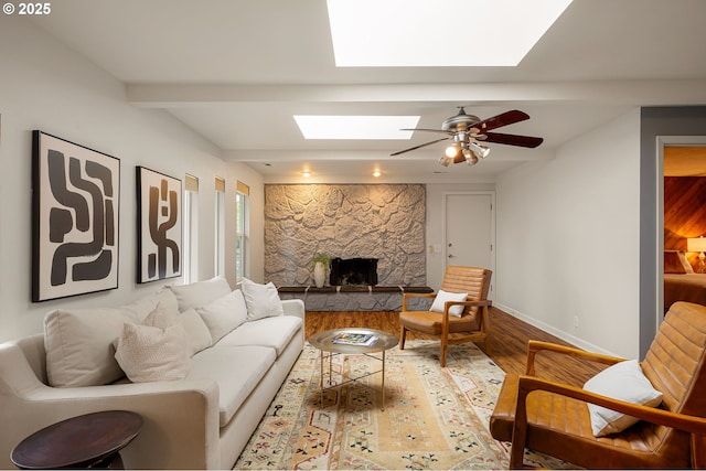 living area featuring a skylight, wood finished floors, a stone fireplace, baseboards, and ceiling fan