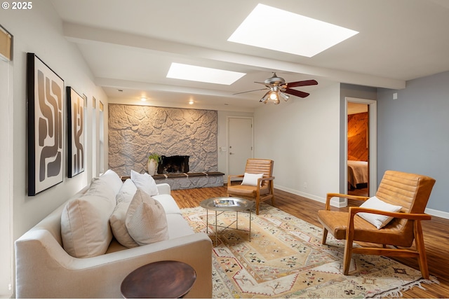 living room with a skylight, wood finished floors, a stone fireplace, baseboards, and a ceiling fan