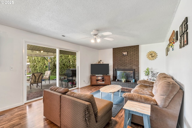 living room featuring ceiling fan, a textured ceiling, wood finished floors, baseboards, and a brick fireplace