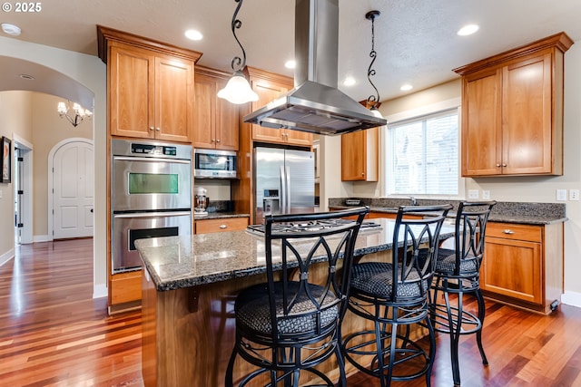 kitchen featuring wood finished floors, a kitchen island, island range hood, dark stone counters, and appliances with stainless steel finishes