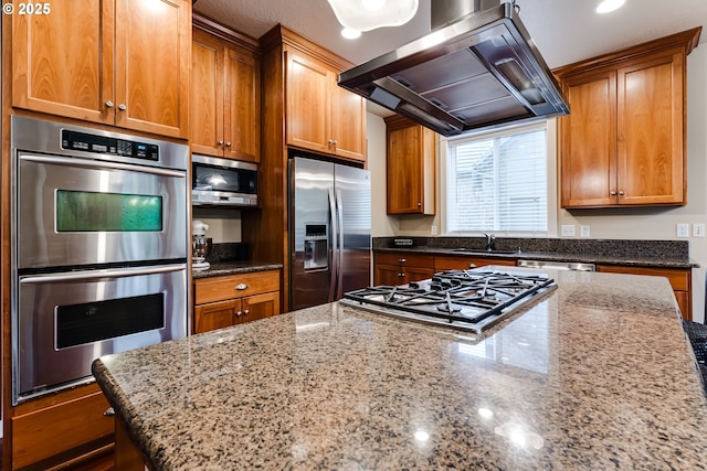 kitchen featuring dark stone counters, appliances with stainless steel finishes, brown cabinets, and wall chimney exhaust hood