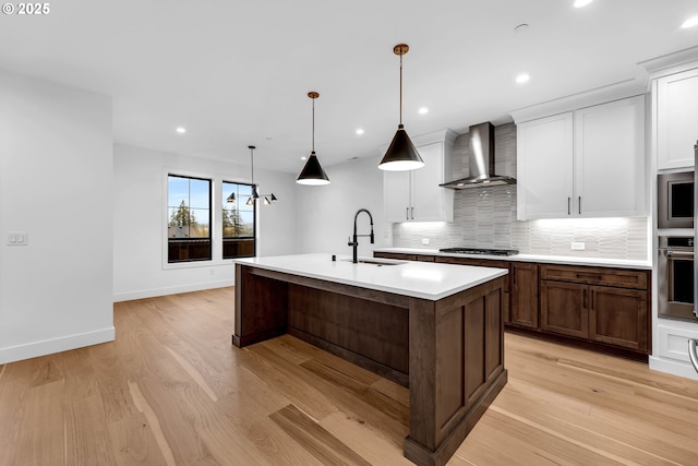 kitchen featuring wall chimney exhaust hood, a sink, light countertops, a center island with sink, and decorative backsplash