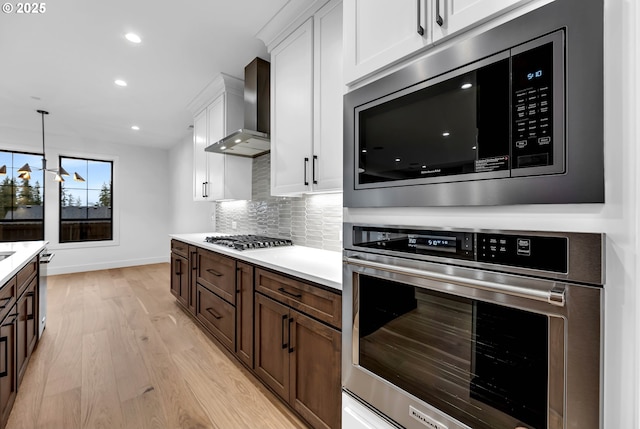 kitchen featuring wall chimney exhaust hood, white cabinetry, stainless steel appliances, light wood finished floors, and baseboards