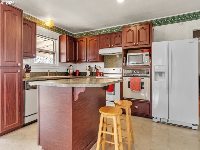 kitchen with a kitchen island, white appliances, under cabinet range hood, a kitchen breakfast bar, and wallpapered walls