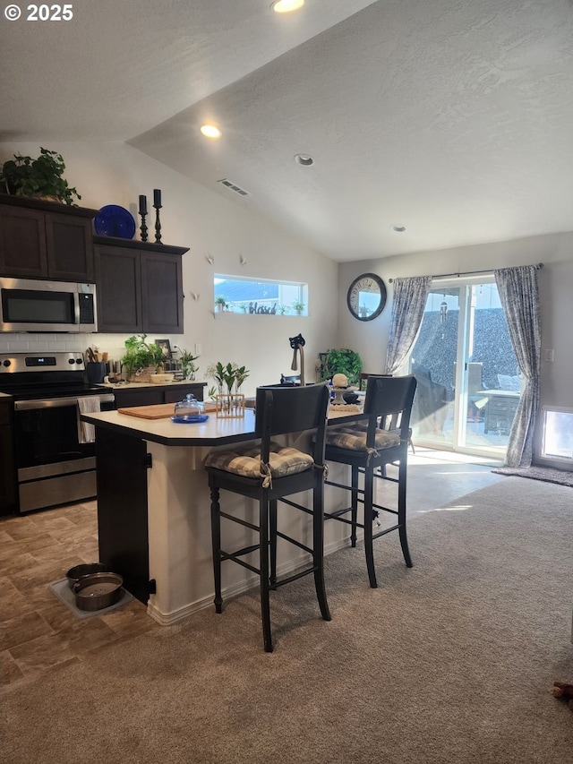 kitchen featuring visible vents, light colored carpet, lofted ceiling, appliances with stainless steel finishes, and a kitchen bar