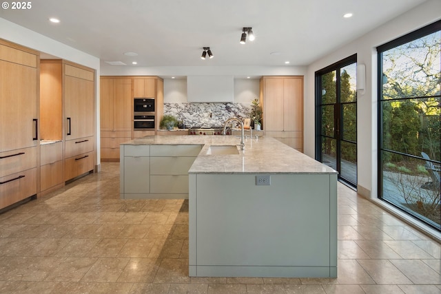 kitchen featuring sink, a large island with sink, light brown cabinets, light stone countertops, and tasteful backsplash