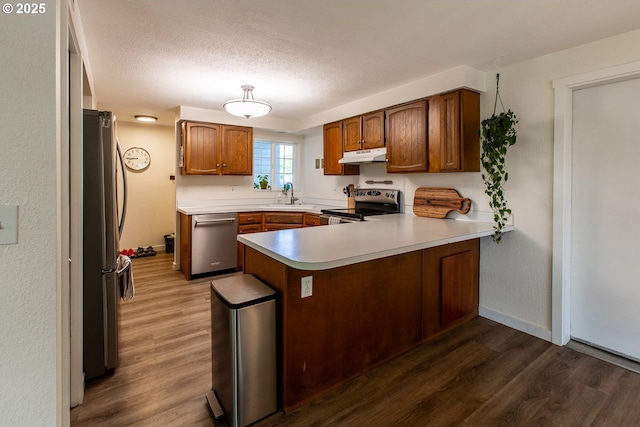 kitchen featuring stainless steel appliances, under cabinet range hood, a peninsula, dark wood-style flooring, and light countertops