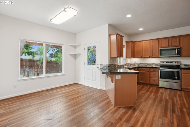kitchen featuring stainless steel appliances, dark countertops, brown cabinetry, a peninsula, and recessed lighting