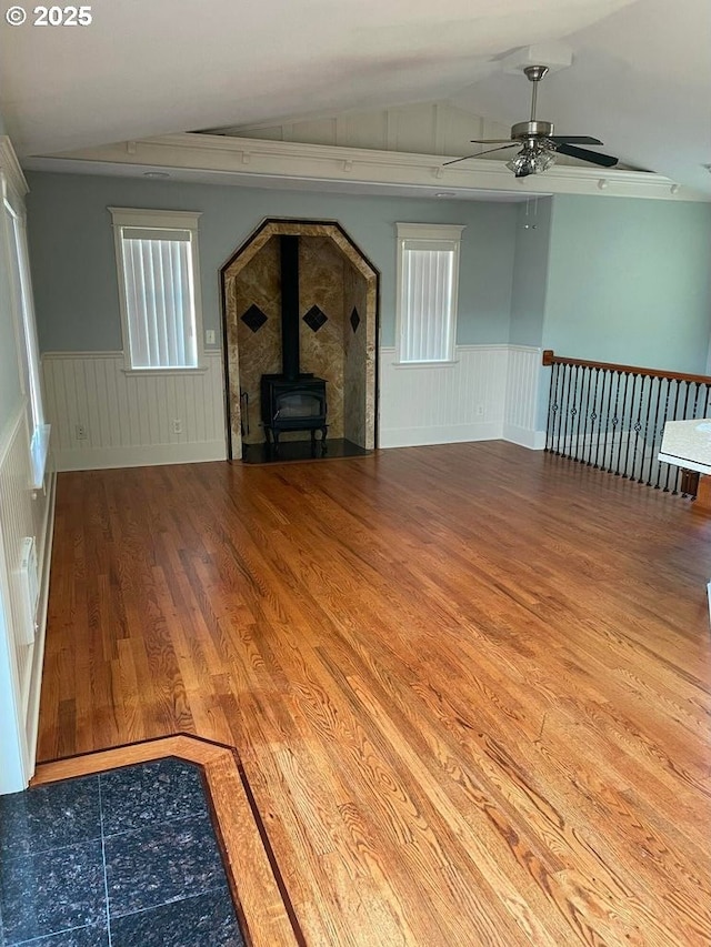unfurnished living room featuring ceiling fan, vaulted ceiling, a wood stove, and a wainscoted wall