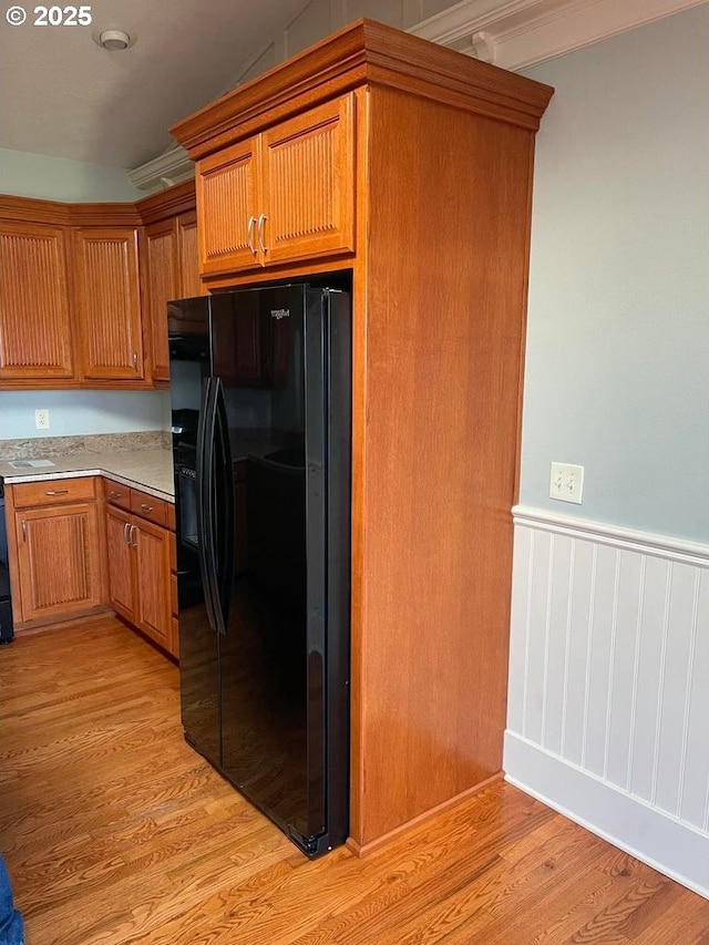 kitchen featuring brown cabinets, black fridge, light wood-style flooring, and a wainscoted wall