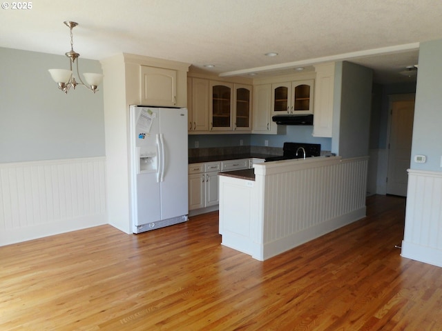 kitchen with white refrigerator with ice dispenser, a wainscoted wall, a peninsula, glass insert cabinets, and under cabinet range hood