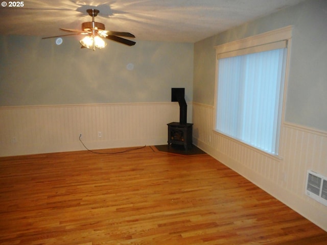 unfurnished living room with a ceiling fan, visible vents, a wood stove, wood finished floors, and a wainscoted wall